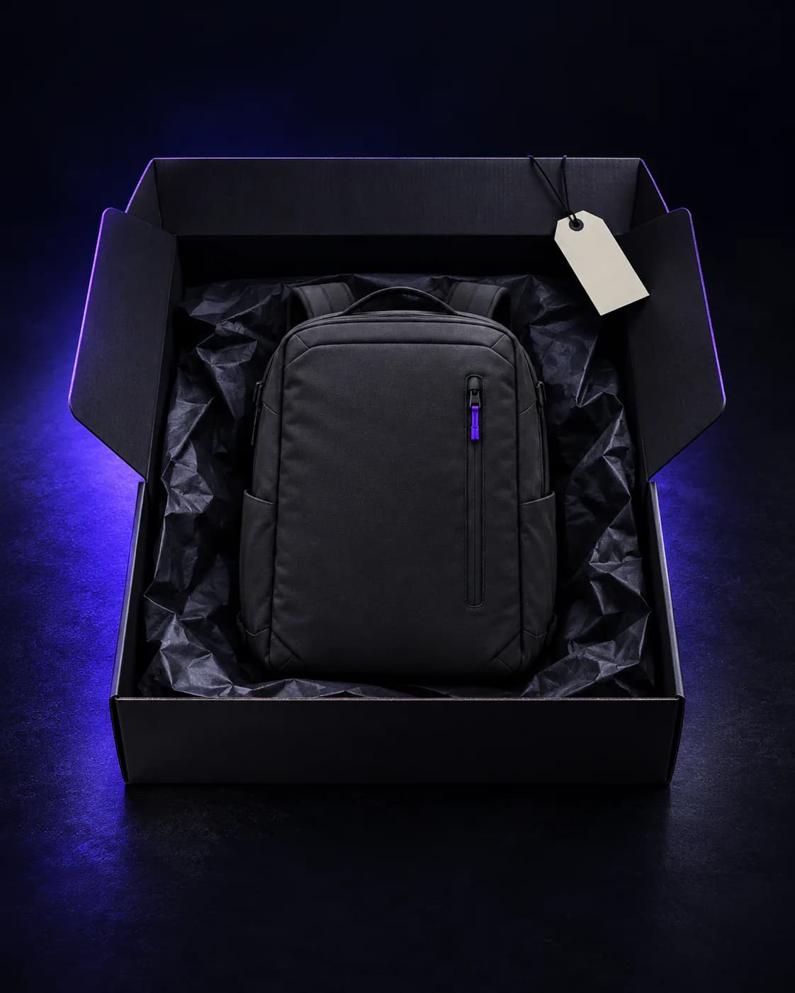 Charcoal backpack folded in black tissue inside an open shipping box, with a blank cream tag on the flap