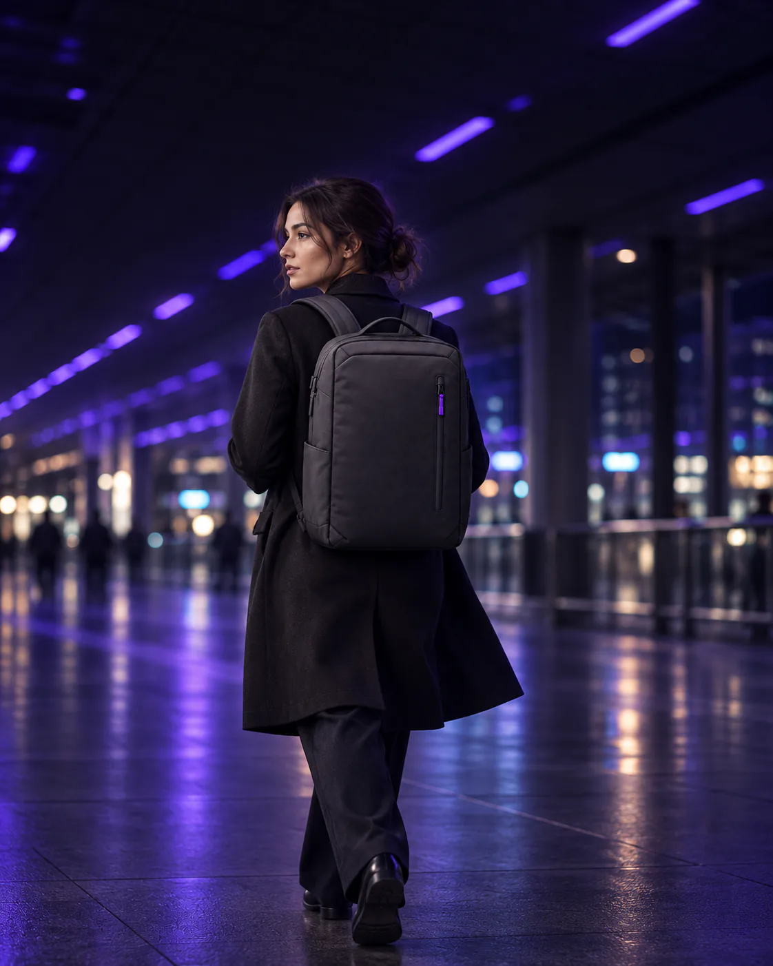 Woman in a dark coat walking through a train station concourse at night, wearing a charcoal backpack
